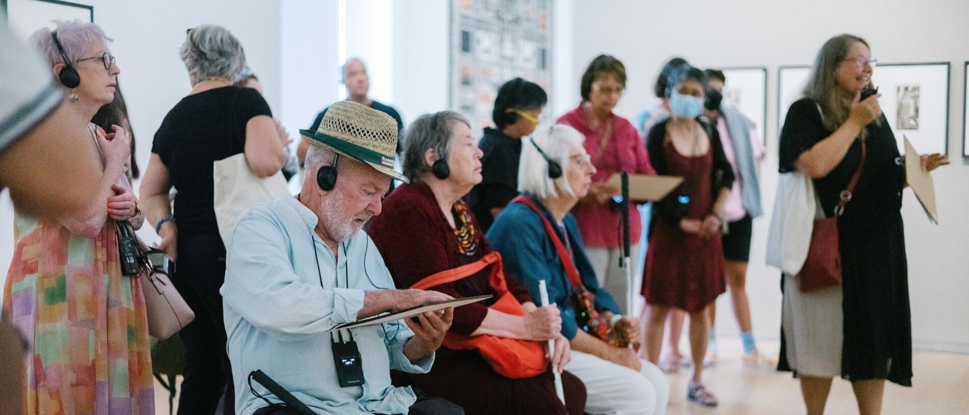 Audio Describer Nicola Owen speaks through a transmitter to a group of Blind people with white canes listening on headphones. Some stand, others sit, one is feeling a touch piece.