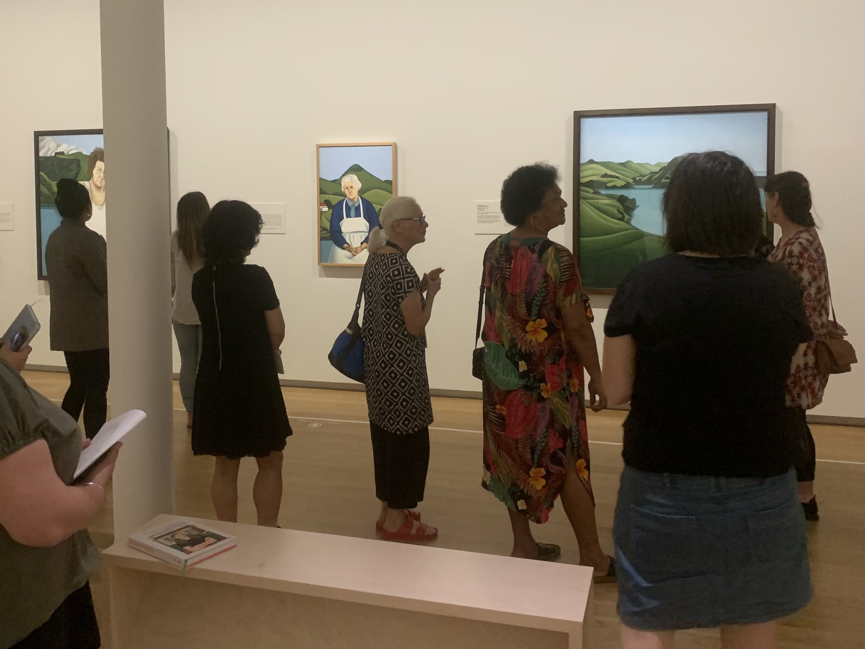 <p>Robin White and kaiako kōrero in&nbsp;<em>Robin White: Te Whanaketanga | Something is Happening Here</em>,&nbsp;Auckland Art Gallery Toi o Tāmaki, November 2022. Details of artworks included (left to right): Robin White, <em>Sam Hunt, Bottle Creek</em>, 1973, oil on canvas, The University of Auckland Art Collection;&nbsp;<em>Florence and Harbour Cone</em>, 1974, oil on canvas, Christchurch Art Gallery Te Puna o Waiwhetū, purchased 1975 with assistance from the Queen Elizabeth II Arts Council;&nbsp;<em>Hooper&#39;s Inlet</em>, 1976, oil on canvas on board, The Fletcher Trust Collection. Image: Kendra Stoner.&nbsp;</p>
