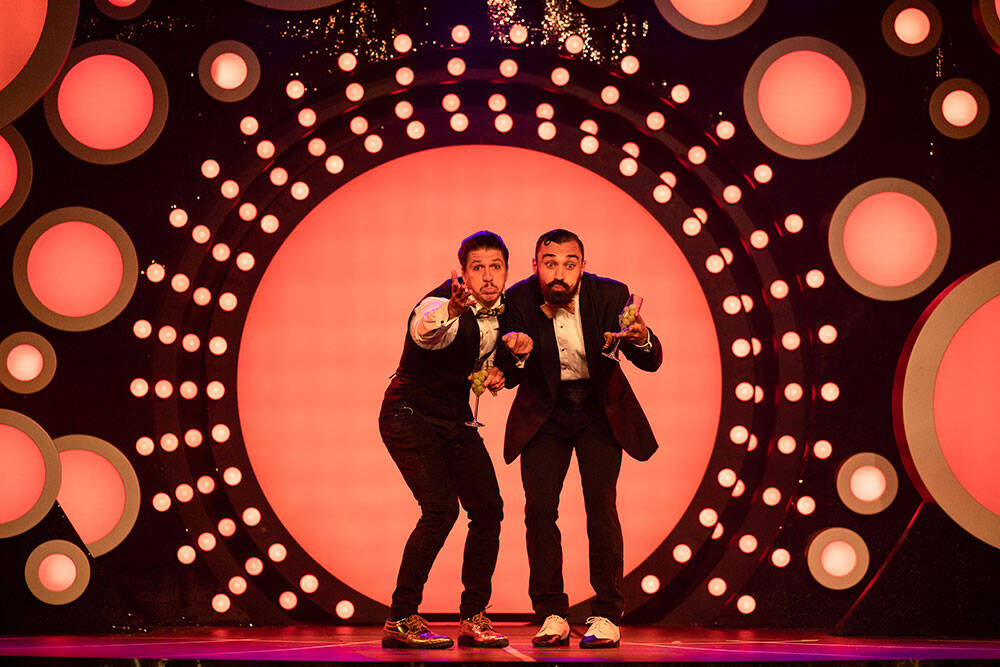 Two performers on stage in suits drinking and laughing
