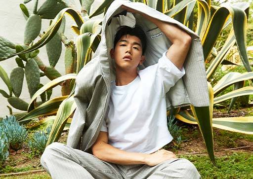 Eric Nam sitting on the grass in front of a flax bush, holding his jacket over his head.
