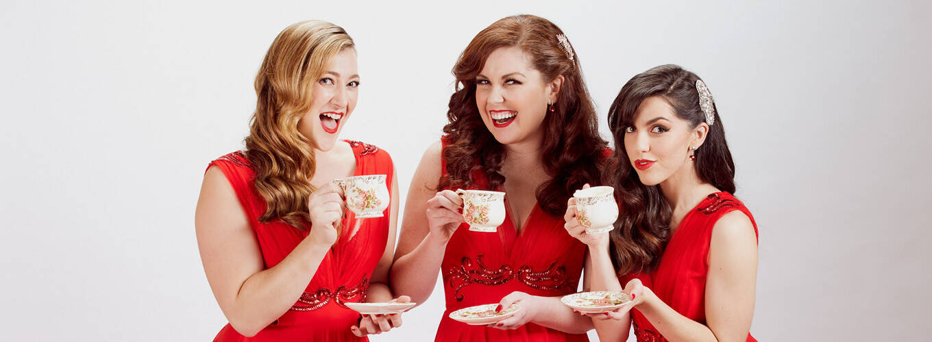 The Madeleines in red dresses, smiling and holding teacups in front of a white background.