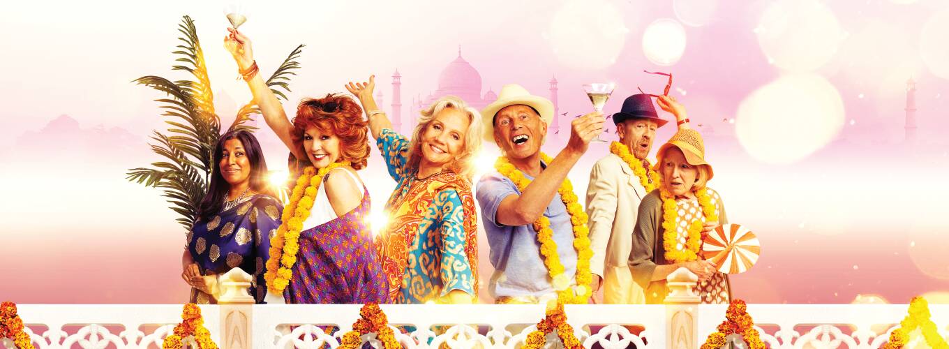 The cast of The Best Exotic Marigold Hotel holding up glasses suggesting "cheers" in front of a pink backdrop with the Taj Mahal faintly in the background.