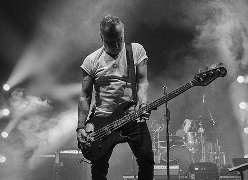Black and white image of Peter Hook standing on a stage with his guitar at hand and looking down.