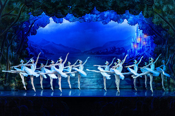 Ballerinas performing in Swan Lake on stage under blue lighting across a lake 