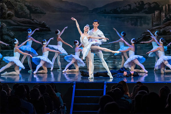 Ballerinas performing in Swan Lake on stage under blue lighting across a lake 