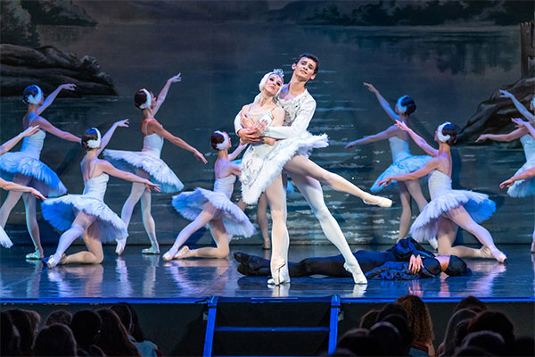 Ballerinas performing in Swan Lake on stage under blue lighting across a lake 