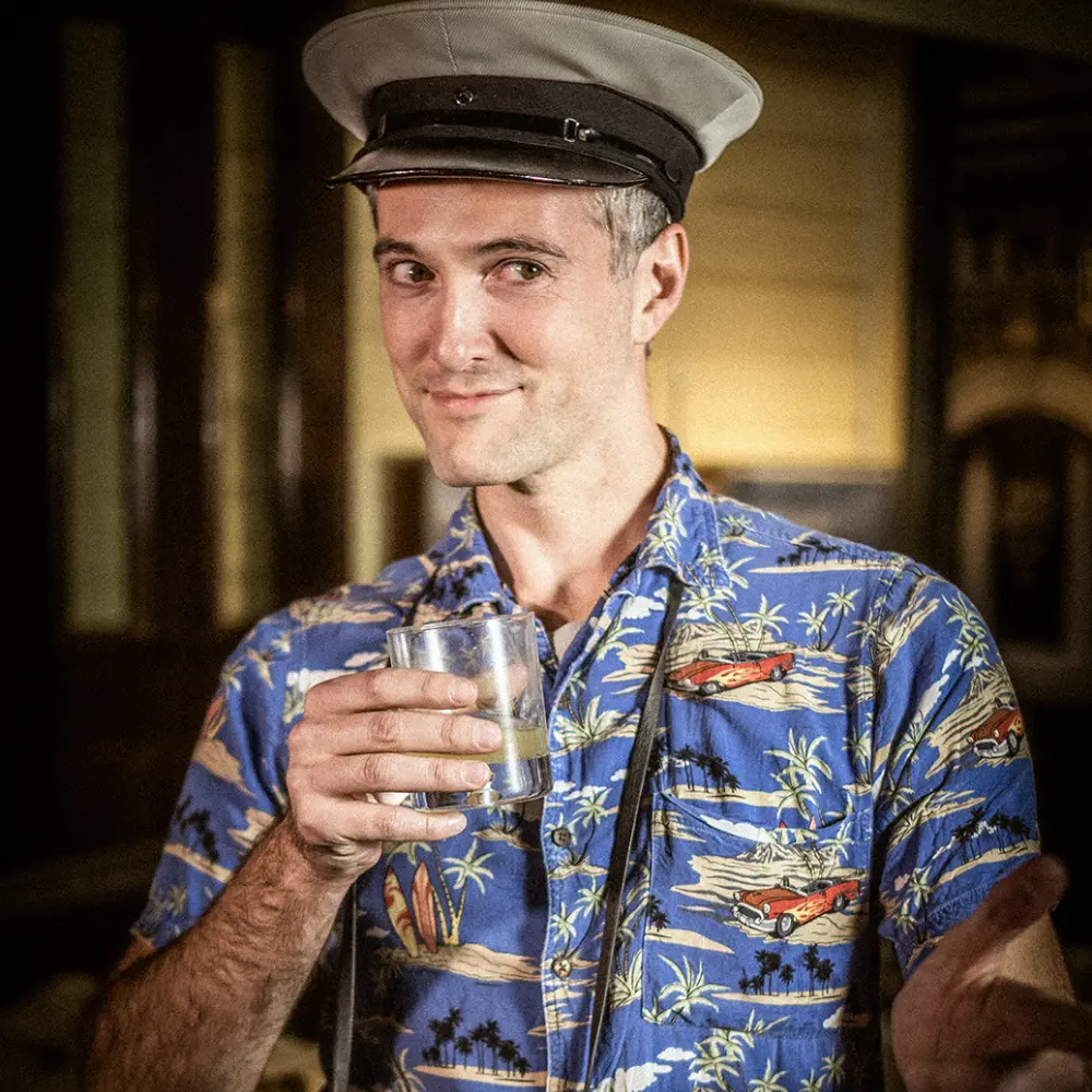 https://cdn.aucklandunlimited.com/maritime/assets/media/while-the-light-lasts-tourist-man-hawaiian-t-shirt-drink-hat.webp