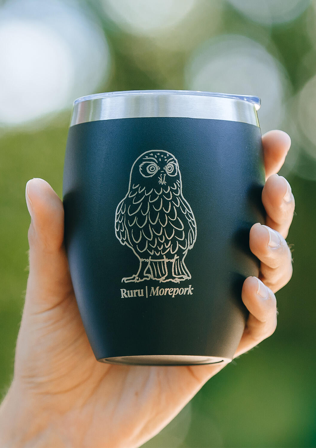 A hand holding an Auckland Zoo black reusable coffee cup featuring an illustration of a Ruru /Morepork